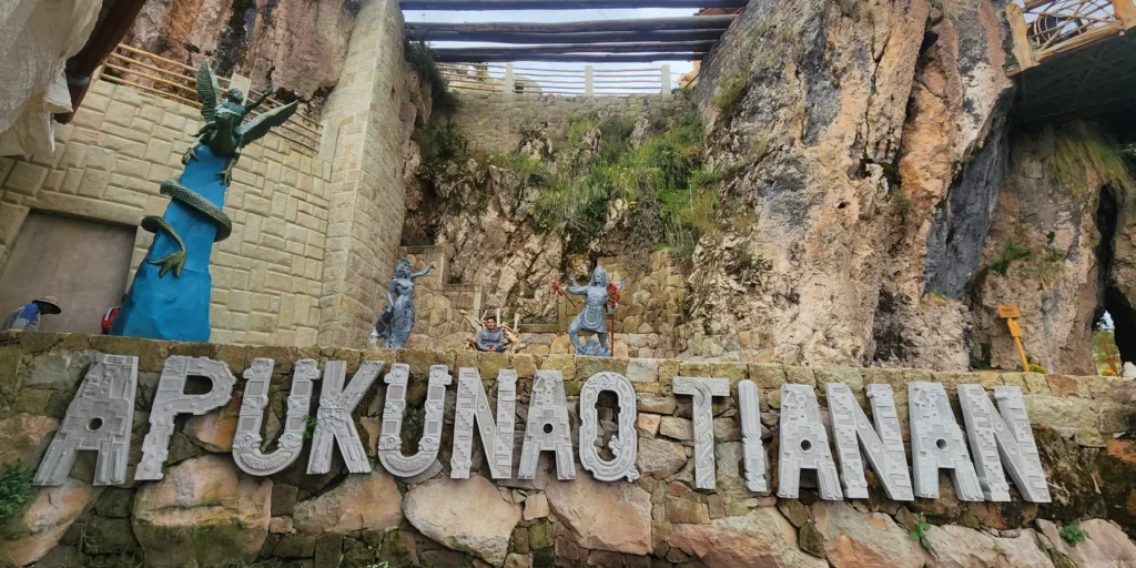 Entrance to the Apukunaq Tianan – The Abode of the Gods site in Cusco, Peru. A large sign with Andean-style lettering can be seen on a natural stone wall. In the background, stone sculptures of gods and mythical beings are carved, including winged figures and warriors with traditional weapons. The site combines architecture inspired by Inca and Quechua culture with spiritual and artistic elements, creating a space dedicated to history, mythology, and the connection with the sacredness of the Andes.