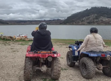 Tourists on ATVs enjoying a view of Lake Huaypo, in Cusco, Peru. Two people wearing helmets observe the lake surrounded by green mountains under a cloudy sky. An outdoor adventure experience near one of the most beautiful lakes in the Sacred Valley.