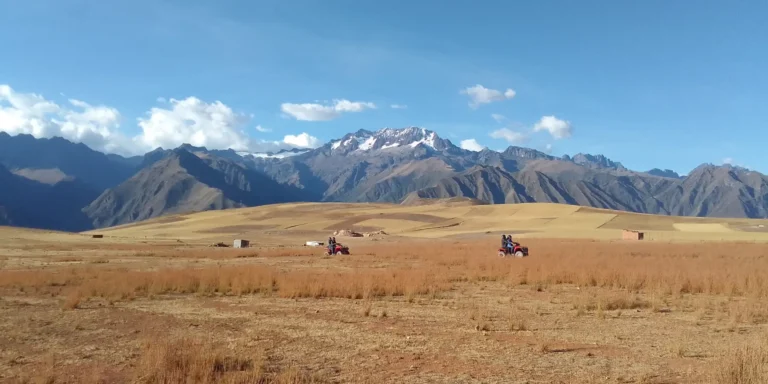 Tourists on ATVs tour a vast Andean countryside near Maras and Moray, in Cusco, Peru. Two vehicles travel across a vast, dry plain under a blue, cloudy sky, with snow-capped mountains in the background. A thrilling adventure experience amidst the stunning landscapes of the Sacred Valley.