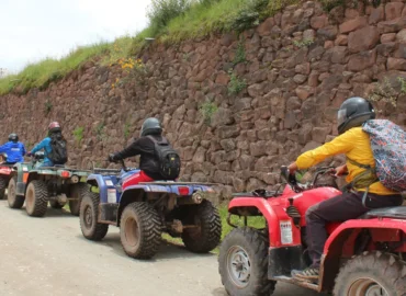 Tourists on ATVs ride along a trail near the Inca ruins of Maras and Moray, in Cusco, Peru. People wearing helmets travel along a dirt road next to an imposing Inca stone wall. An adventure experience that combines nature, history, and excitement in the Sacred Valley.