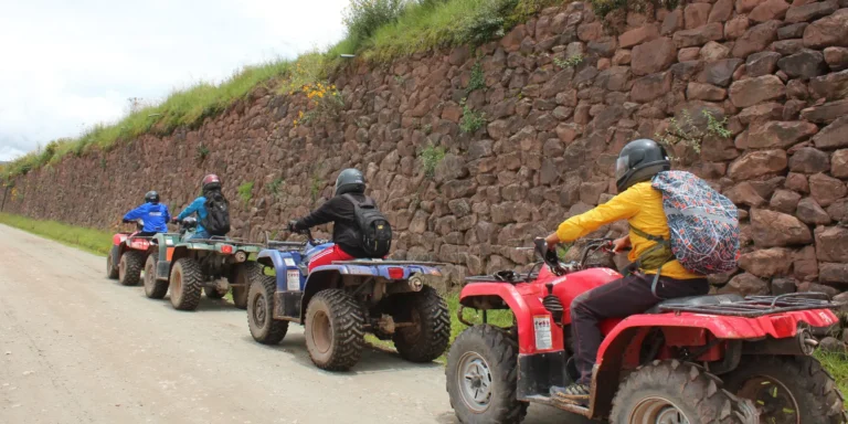 Tourists on ATVs ride along a trail near the Inca ruins of Maras and Moray, in Cusco, Peru. People wearing helmets travel along a dirt road next to an imposing Inca stone wall. An adventure experience that combines nature, history, and excitement in the Sacred Valley.