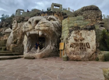 Entrance to the Apukunaq Tianan – Abode of the Gods site in Cusco, Peru. A large stone sculpture carved in the shape of a lion's head with an open mouth can be seen, with a person sitting inside. Beside it, a rock with the name "Apukunaq Tianan – Abode of the Gods" engraved in relief, along with Andean symbols. In the background, stone structures with stairs and a carved figure representing a mythological being. This site combines Andean art, culture, and spirituality, inspired by the ancestral traditions of the indigenous peoples of the Andes.