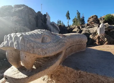 Stone sculpture in the shape of a serpent's head in the Valley of the Goblins, Cusco, Peru. The carving shows intricate details in its scaly skin and deep-set eyes, representing an important mythical figure in Andean culture. In the background, other stone sculptures, such as a large ear of corn and animal forms, can be seen on natural rocks under a clear blue sky. The site is a cultural and tourist space that celebrates the legends, symbols, and ancestral traditions of the indigenous peoples of the Andes.