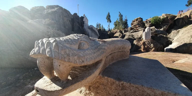 Stone sculpture in the shape of a serpent's head in the Valley of the Goblins, Cusco, Peru. The carving shows intricate details in its scaly skin and deep-set eyes, representing an important mythical figure in Andean culture. In the background, other stone sculptures, such as a large ear of corn and animal forms, can be seen on natural rocks under a clear blue sky. The site is a cultural and tourist space that celebrates the legends, symbols, and ancestral traditions of the indigenous peoples of the Andes.