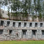 Themed building in the Valley of the Goblins, Cusco, Peru. The structure is built with Inca-style carved stones, with rectangular windows and symbolic details such as geometric motifs and ancient vessels on the walls. It is surrounded by green grass and tall trees under a cloudy sky. This place combines art, culture, and Andean legends, offering visitors an immersive experience in the history and mythology of the indigenous peoples of the Andes.