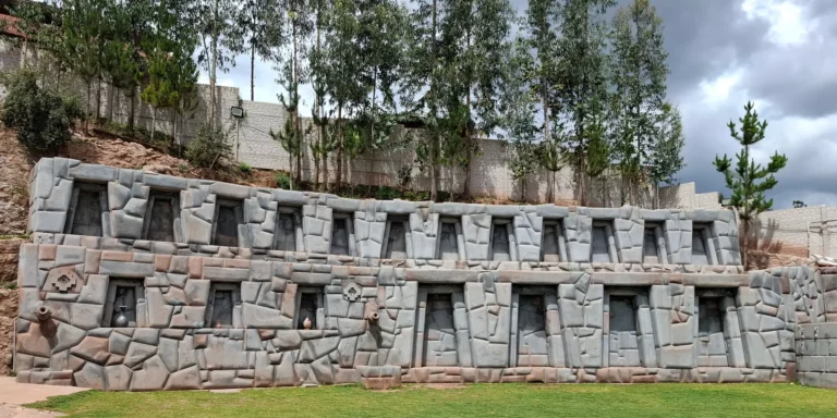 Themed building in the Valley of the Goblins, Cusco, Peru. The structure is built with Inca-style carved stones, with rectangular windows and symbolic details such as geometric motifs and ancient vessels on the walls. It is surrounded by green grass and tall trees under a cloudy sky. This place combines art, culture, and Andean legends, offering visitors an immersive experience in the history and mythology of the indigenous peoples of the Andes.