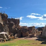Valley of the Goblins theme park, Cusco, Peru. Large rock formations with stone sculptures depicting human figures, mythical figures, and cultural symbols can be seen. Some are located on stairs or rocks, while others blend into the natural landscape. Wooden benches, umbrellas, and visitors explore the area under a clear blue sky with white clouds. The site celebrates Andean mythology, ancestral traditions, and the connection between nature and the spirit of the Quechua people.