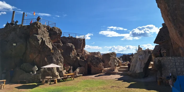Valley of the Goblins theme park, Cusco, Peru. Large rock formations with stone sculptures depicting human figures, mythical figures, and cultural symbols can be seen. Some are located on stairs or rocks, while others blend into the natural landscape. Wooden benches, umbrellas, and visitors explore the area under a clear blue sky with white clouds. The site celebrates Andean mythology, ancestral traditions, and the connection between nature and the spirit of the Quechua people.