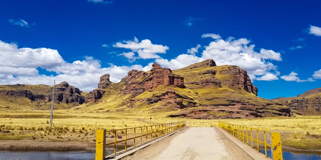 Puente sobre un río en el Cañón de Tinajani, Perú. Vista panorámica de formaciones rocosas rojizas y llanuras andinas bajo un cielo azul. Destino natural en Puno, ideal para caminatas y explorar las maravillas geológicas del altiplano. Escenario perfecto para viajeros que buscan paisajes únicos en el sur del Perú