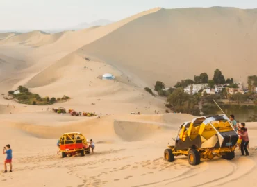 Turistas aproveitam uma aventura de buggy nas dunas de Huacachina, Peru. Veículos amarelos e vermelhos percorrem as impressionantes dunas sob um céu limpo, enquanto as pessoas caminham e tiram fotos. Ao fundo, avista-se um oásis com palmeiras e água, um lugar mágico para vivenciar emoções no deserto.