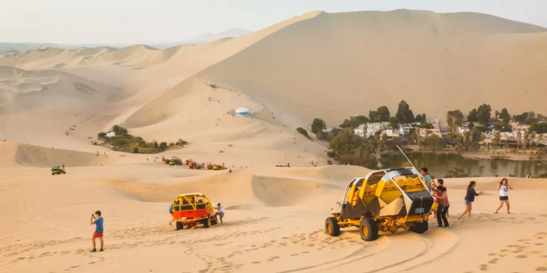 Turistas aproveitam uma aventura de buggy nas dunas de Huacachina, Peru. Veículos amarelos e vermelhos percorrem as impressionantes dunas sob um céu limpo, enquanto as pessoas caminham e tiram fotos. Ao fundo, avista-se um oásis com palmeiras e água, um lugar mágico para vivenciar emoções no deserto.