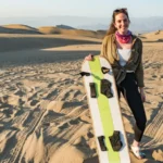 Mulher sorridente segurando uma prancha de sandboard nas dunas de Huacachina, Peru. Ela está em pé sobre areia dourada com imponentes montanhas de areia ao fundo, sob um céu limpo. Uma experiência divertida e emocionante de esportes radicais no deserto mais famoso do país.