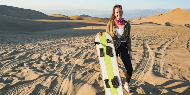 Mulher sorridente segurando uma prancha de sandboard nas dunas de Huacachina, Peru. Ela está em pé sobre areia dourada com imponentes montanhas de areia ao fundo, sob um céu limpo. Uma experiência divertida e emocionante de esportes radicais no deserto mais famoso do país.