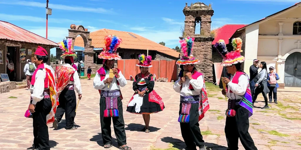Música tradicional en Taquile, isla del Lago Titicaca, Perú. Hombres vestidos con trajes típicos andinos tocan flautas de pan mientras mujeres lucen faldas bordadas y sombreros coloridos. Escena cultural en la plaza principal de Taquile, donde se conserva la tradición ancestral de tejido y música. Destino turístico emblemático en el sur del Perú, cerca de Puno."