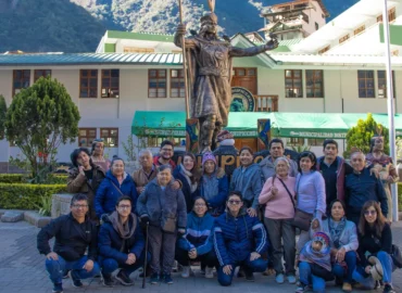 Um grupo de turistas posa em frente à estátua inca no centro de Aguas Calientes, Peru. A imagem mostra pessoas de todas as idades sorrindo ao lado de uma escultura histórica, com montanhas verdejantes e construções típicas locais ao fundo. Um momento feliz durante uma viagem a Machu Picchu.