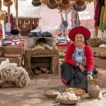 Indigenous community in Chinchero, Cusco, Peru. Women dressed in traditional Andean costumes—red chullos, black skirts, and embroidered jackets—work with alpaca wool at an artisan market. Around them are colorful balls of wool, textiles, and traditional utensils. This is a place where Quechua culture and crafts are preserved in the Sacred Valley.