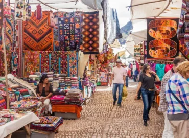 Pisac artisan market in Cusco, Peru. Stalls filled with colorful textiles, blankets, bags, and traditional souvenirs with Andean designs. Tourists walk along cobblestone streets while local women sell their wares under awnings. A vibrant place to learn about the culture and crafts of indigenous peoples.