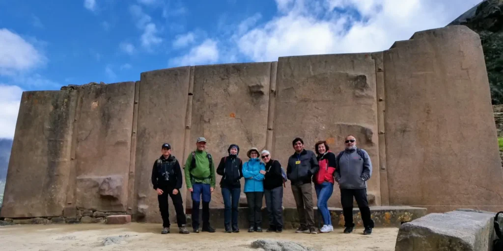 A group of tourists posing in front of the Temple of the Sun in Ollantaytambo, Cusco, Peru. Large carved stone blocks form part of this impressive Inca temple, built to observe the movements of the sun. In the background, a blue sky with clouds and green mountains demonstrate the advanced architecture and power of the Inca civilization.