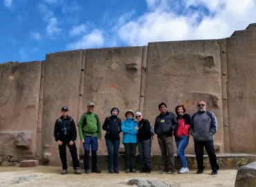 A group of tourists posing in front of the Temple of the Sun in Ollantaytambo, Cusco, Peru. Large carved stone blocks form part of this impressive Inca temple, built to observe the movements of the sun. In the background, a blue sky with clouds and green mountains demonstrate the advanced architecture and power of the Inca civilization.