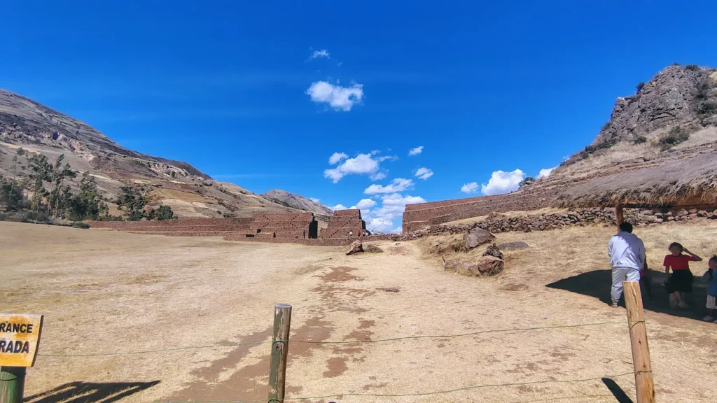 Estructura piramidal de piedra en el sitio arqueológico de Pikillaqta, Cusco, Perú. Se observa una plataforma escalonada de piedra rojiza con bloques irregulares, típica de la arquitectura Wari. Una persona se encuentra de pie en la cima, destacando la escala del monumento. Alrededor, otras ruinas antiguas, colinas verdes y un cielo parcialmente nublado. Pikillaqta fue una importante ciudad preincaica que muestra el desarrollo político, religioso y urbano de la cultura Wari antes del auge incaico.