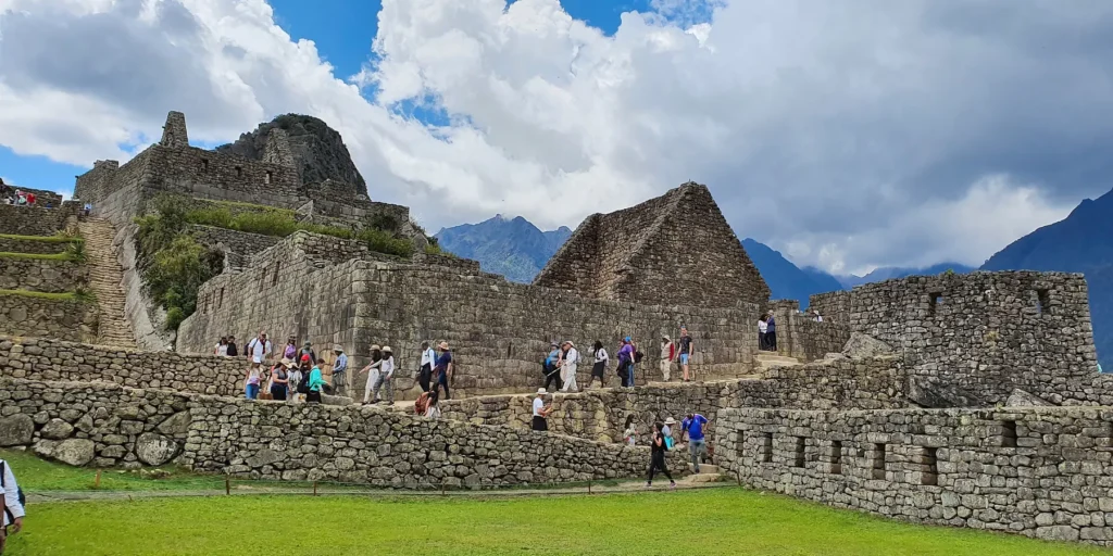 Turistas exploram as antigas estruturas de pedra de Machu Picchu, no Peru. Muralhas e edifícios incas bem preservados erguem-se entre montanhas verdejantes sob um céu azul nublado. Com vistas deslumbrantes da arquitetura inca e da paisagem andina, é um dos locais mais visitados do mundo.