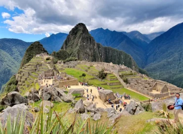 Vista panorâmica de Machu Picchu, Peru, com suas antigas ruínas incas em meio a montanhas verdejantes e um céu azul. Turistas passeiam pelos terraços e ruas de paralelepípedos, admirando a deslumbrante paisagem andina. Um dos locais mais famosos do mundo e Patrimônio Mundial.
