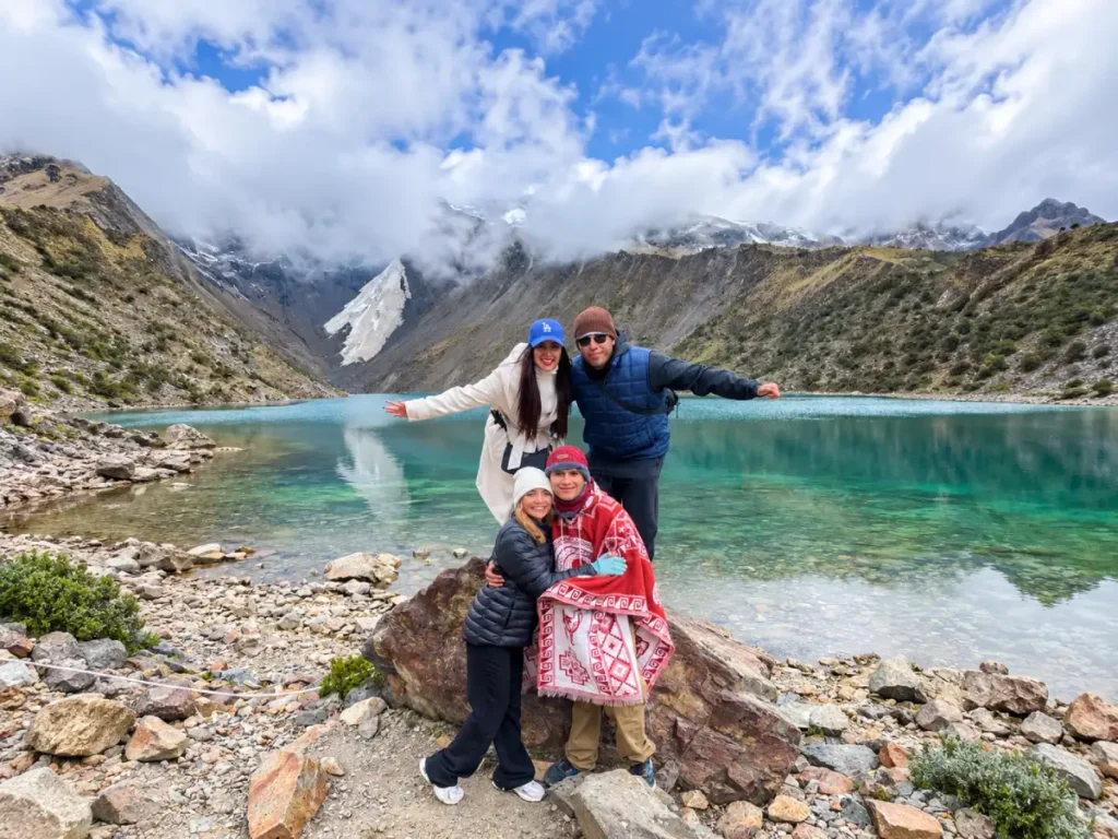Grupo de turistas posando frente a la Laguna Humantay con montañas nevadas y cielo despejado en Cusco, Perú